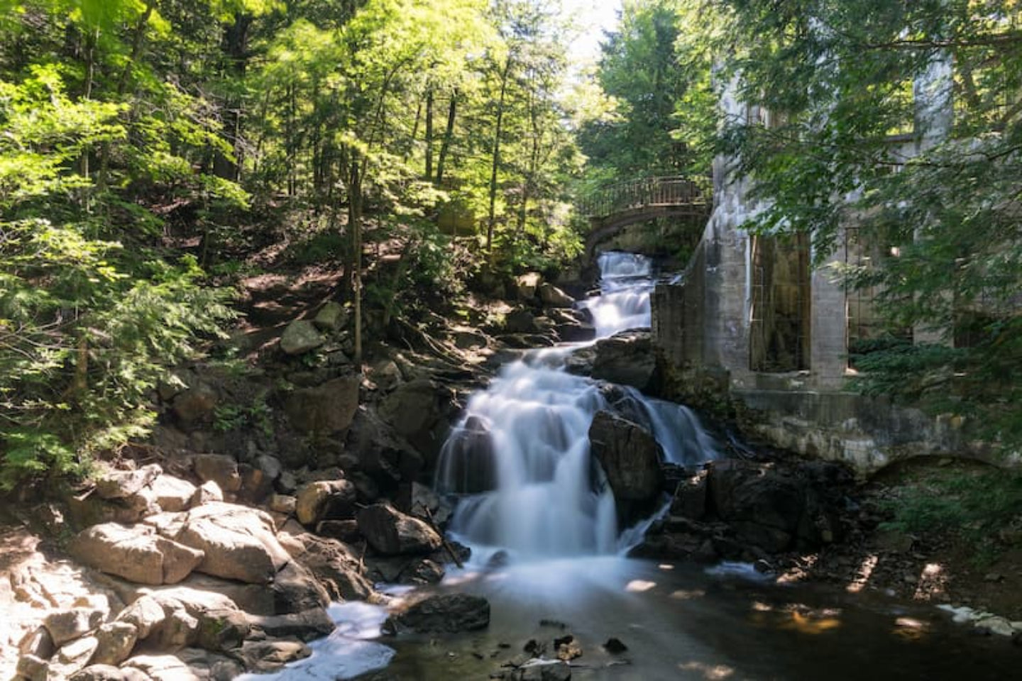 Waterfall in a forest with a stone structure, surrounded by green trees.